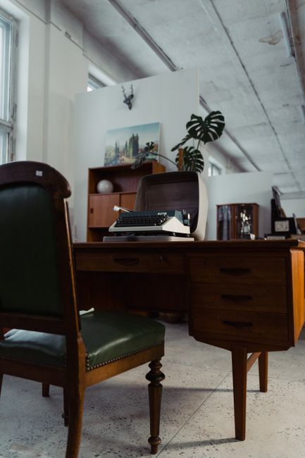 A cluttered office desk with a dark wooden surface, situated in a room with wooden-paneled walls and a window with vertical blinds. The desk holds a vintage beige typewriter partially visible on the l