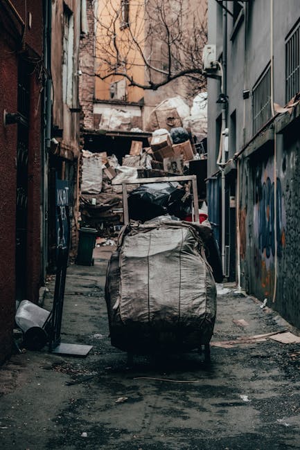A person wearing a plaid shirt with yellow, grey, and blue tones, along with grey trousers secured with a brown belt, is holding open a large black rubbish bag lined with a grey or metallic inner laye