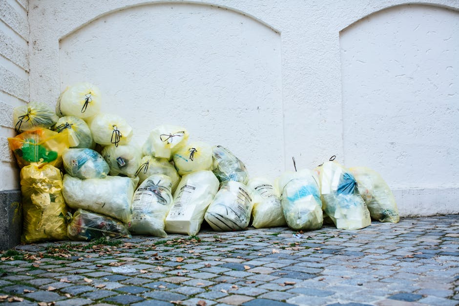 An outdoor scene showing a collection of overflowing rubbish bins and scattered waste on a paved area in front of a commercial building. The waste includes crumpled paper, cardboard boxes, plastic pac