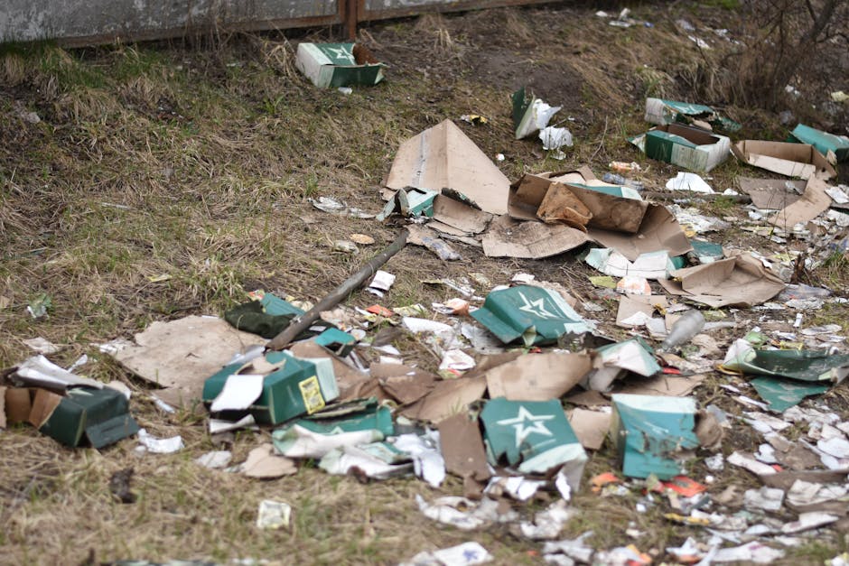 A section of ground covered with discarded packaging materials and rubbish, predominantly composed of flattened cardboard boxes with green and white branding, some torn and crumpled, scattered across