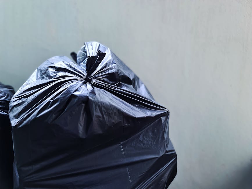 A woman stands on a narrow residential sidewalk next to a row of black wheelie bins used for waste disposal, with some bins labeled with white graffiti. The bins are lined up along brick houses with w