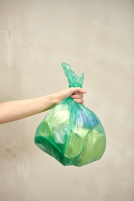 A person's hand and forearm are visible, holding a green plastic rubbish bag by its twisted top. The bag appears to contain flexible waste, with some areas slightly bulging and showing hints of blue a