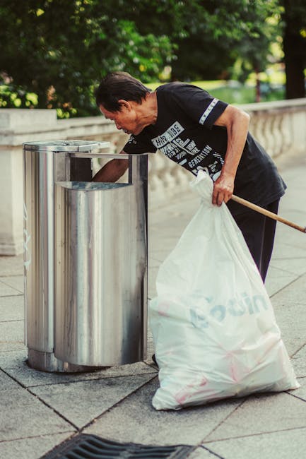 A person's hand and forearm are visible, holding a green plastic rubbish bag by its twisted top. The bag appears to contain flexible waste, with some areas slightly bulging and showing hints of blue a