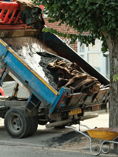 A small burgundy flatbed truck parked on an urban street in front of a modern office building with large glass windows and a concrete facade. The truck's bed is loaded with various types of waste mate